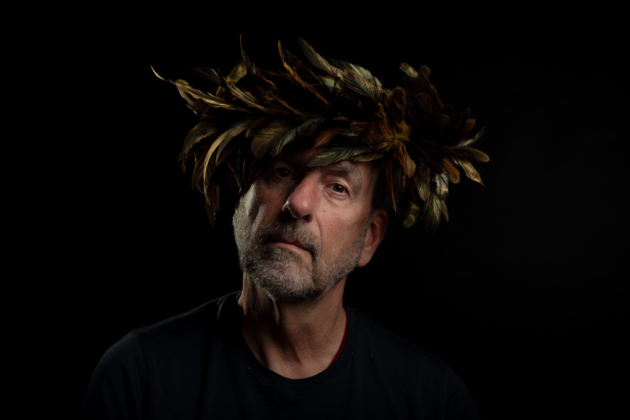 A grey-bearded sitter in a black shirt tilts their head beneath a golden feather crown against a black background.