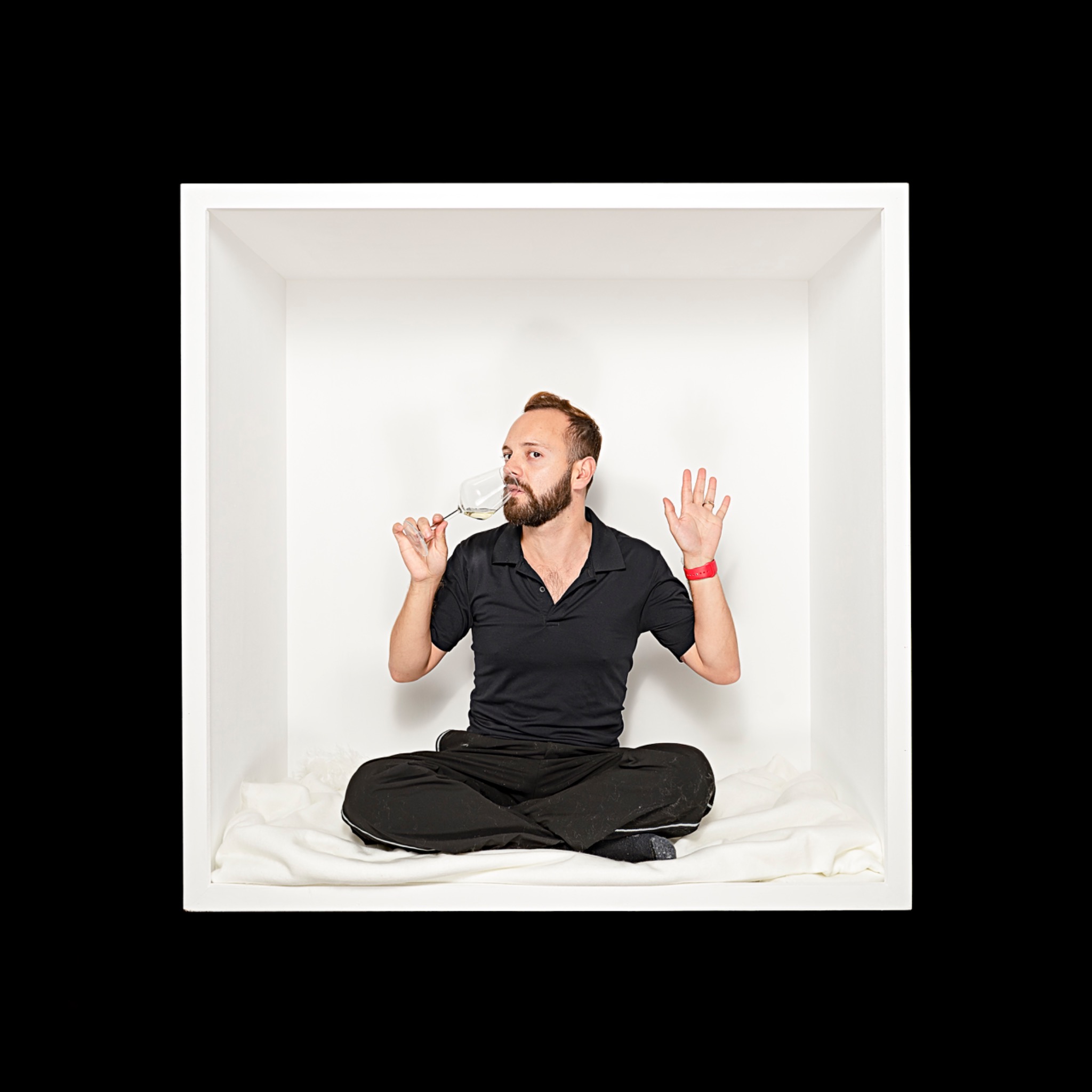 A bearded sitter in black sits cross-legged, drinking from a wine glass with an open hand raised.