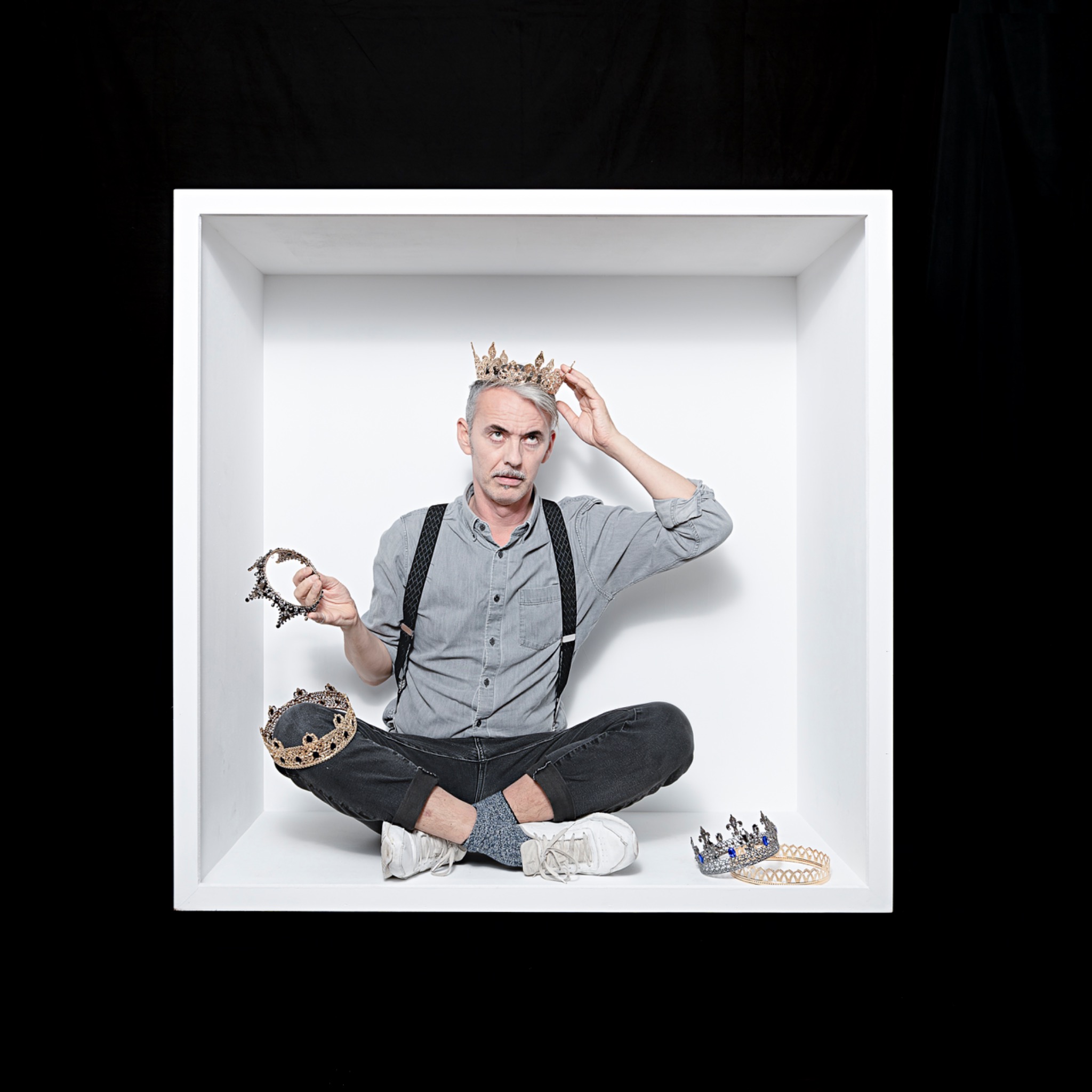 The grey-haired sitter sits cross-legged among crowns, touching one on their head and holding another.