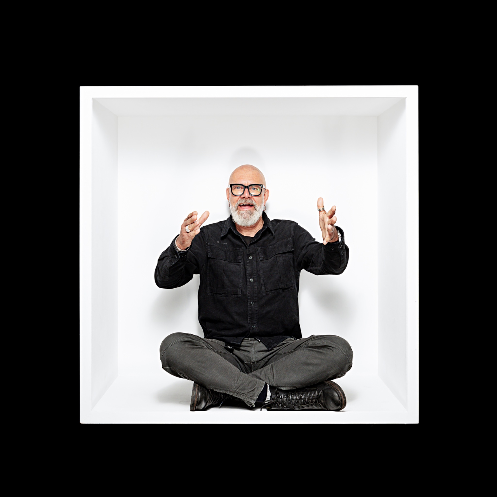 A grey-bearded sitter in black sits cross-legged in the white box, with both hands raised in front.