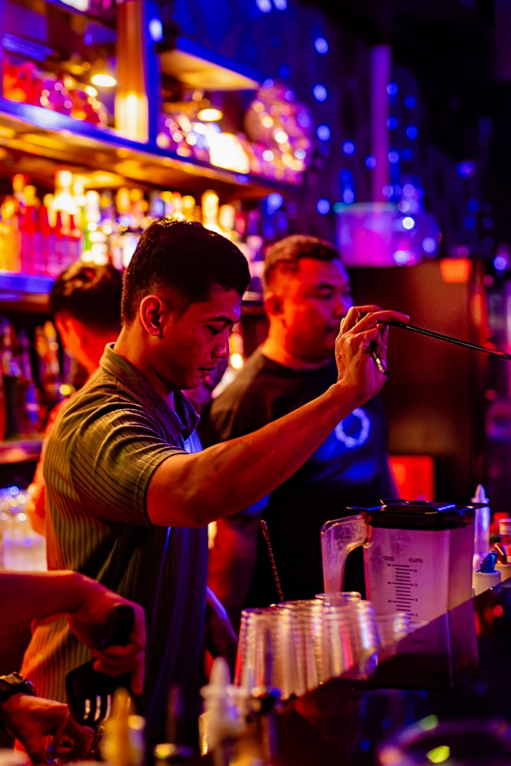 A bartender works in front of glowing bottles, surrounded by gold, blue and magenta light.