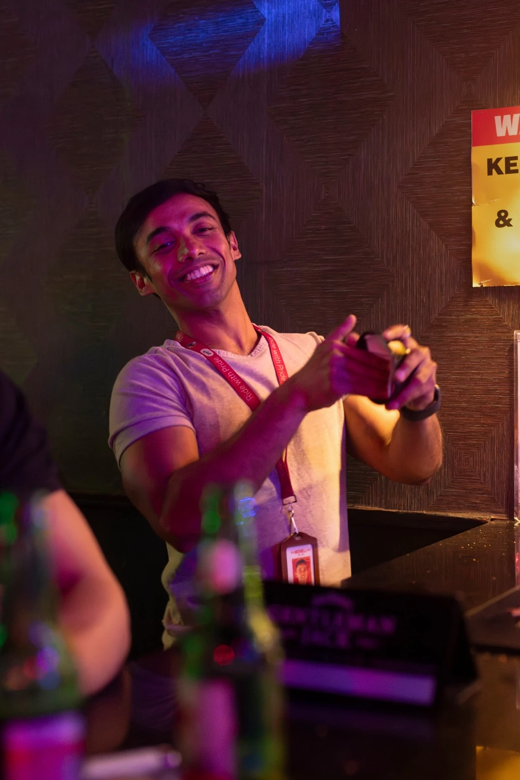 A smiling person behind the bar meets the camera in pink and amber light.
