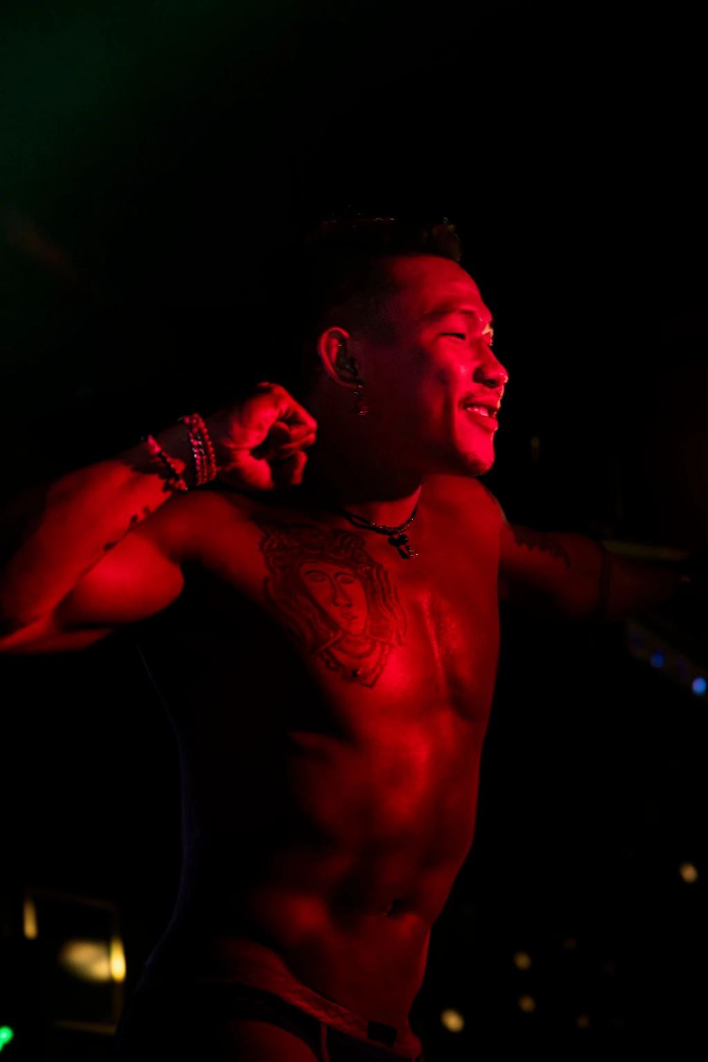 A smiling tattooed dancer raises an arm, illuminated in deep red against a dark room.