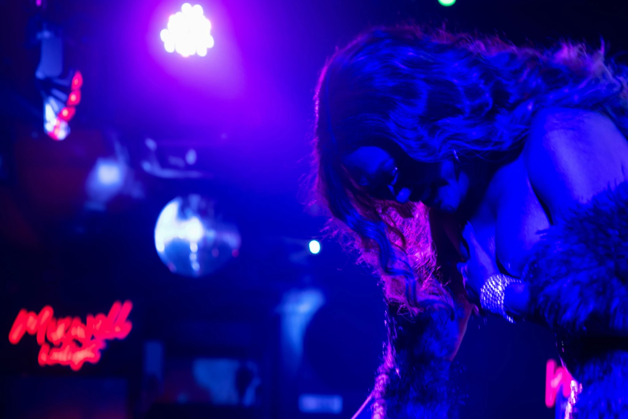 A feathered performer lowers their gaze in blue light beside the red Mixwell neon sign.