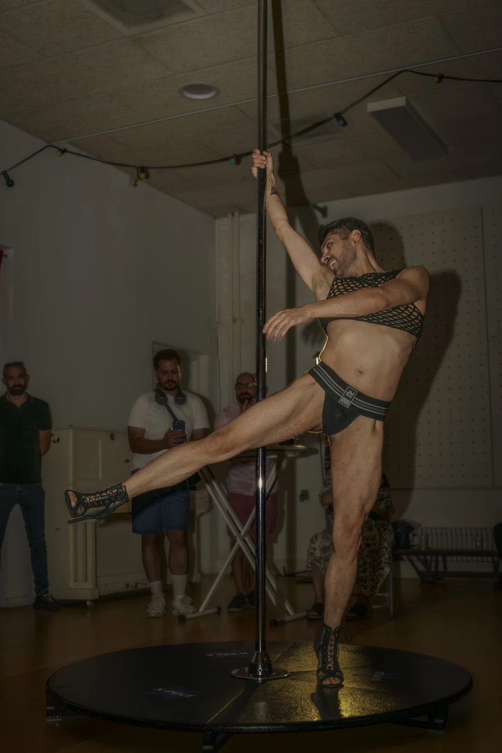 A performer extends one leg beside a pole as guests watch from the edge of the room.
