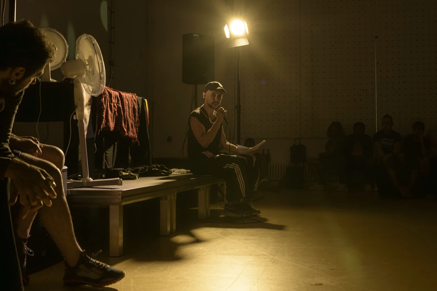 A seated speaker holds a microphone and a sheet of paper under a warm spotlight, with listeners around the room.