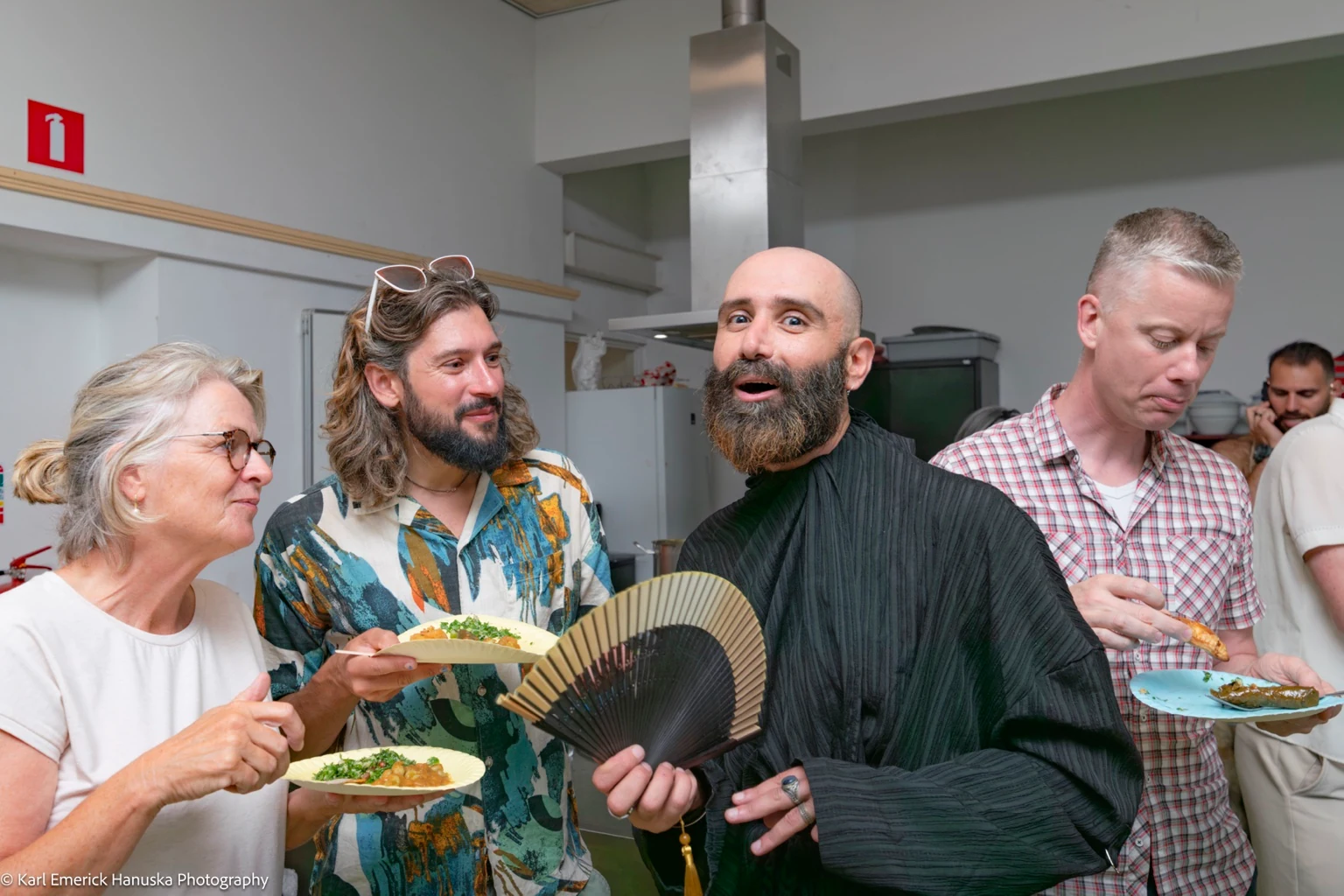 Guests share food and conversation in the kitchen as a person in black holds an open fan.