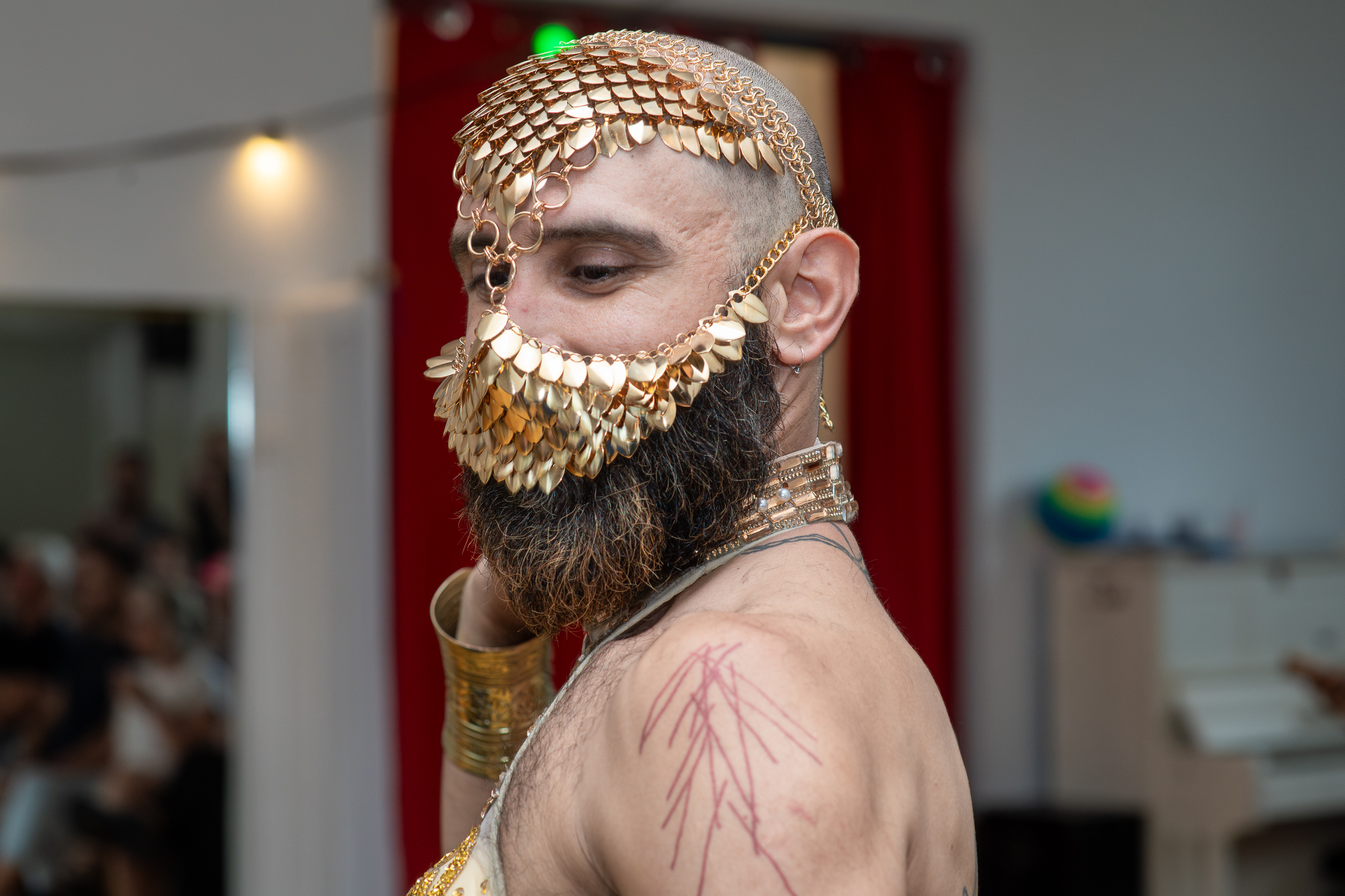Close portrait of a bearded person wearing a gold headdress and beaded face covering, looking over an exposed shoulder.