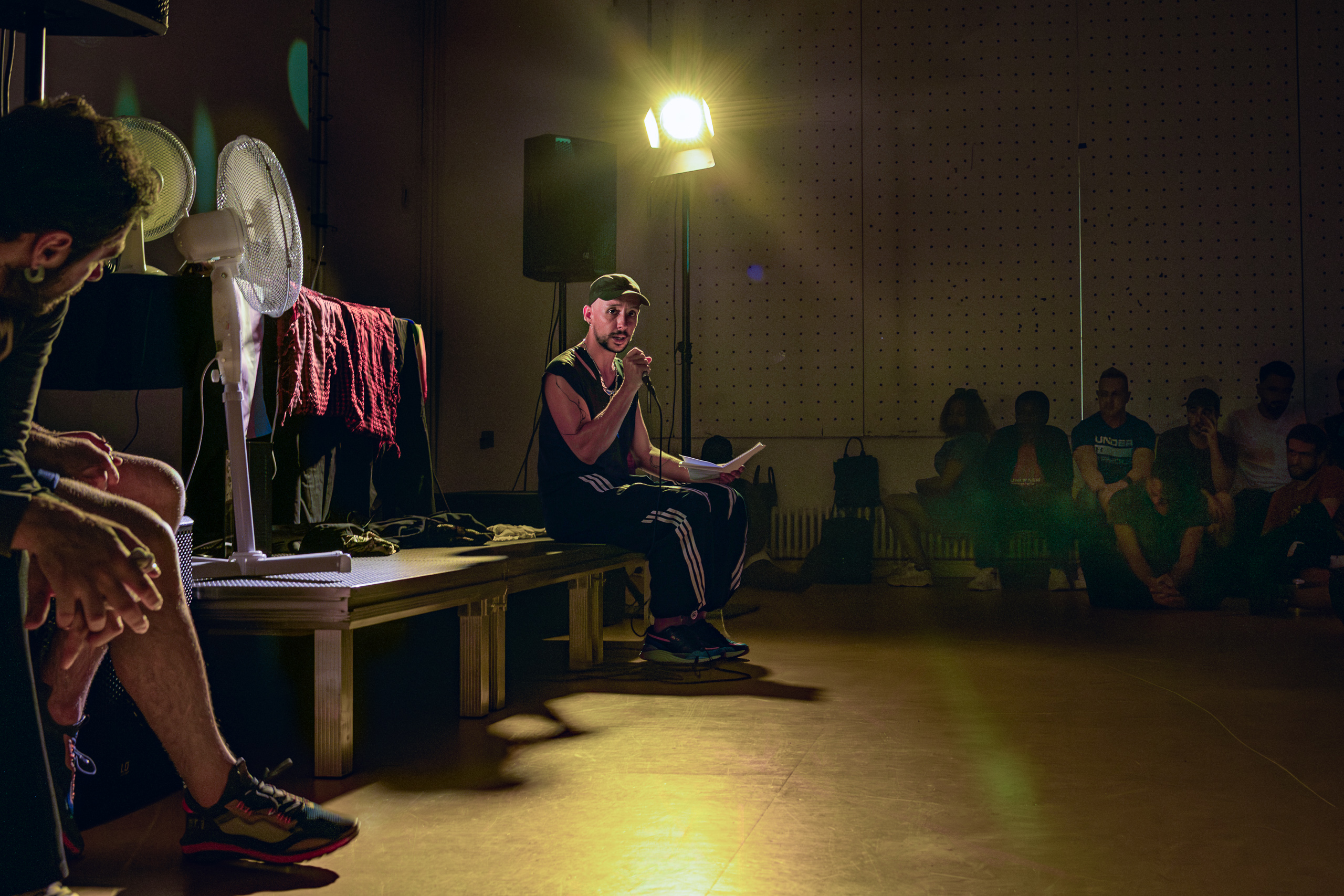 A seated speaker holds a microphone beneath a single light, with listeners around the room.