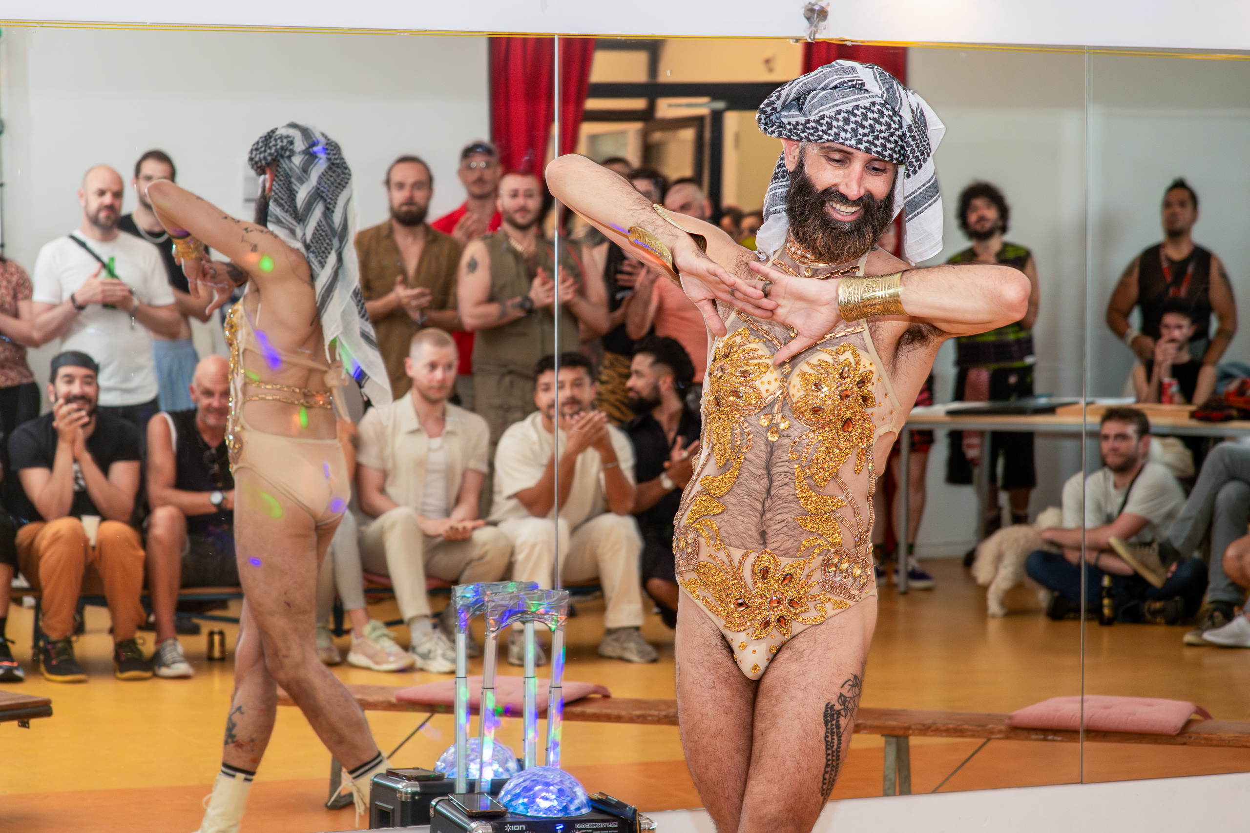 A performer in a gold costume crosses their hands, with the body reflected in a mirror.