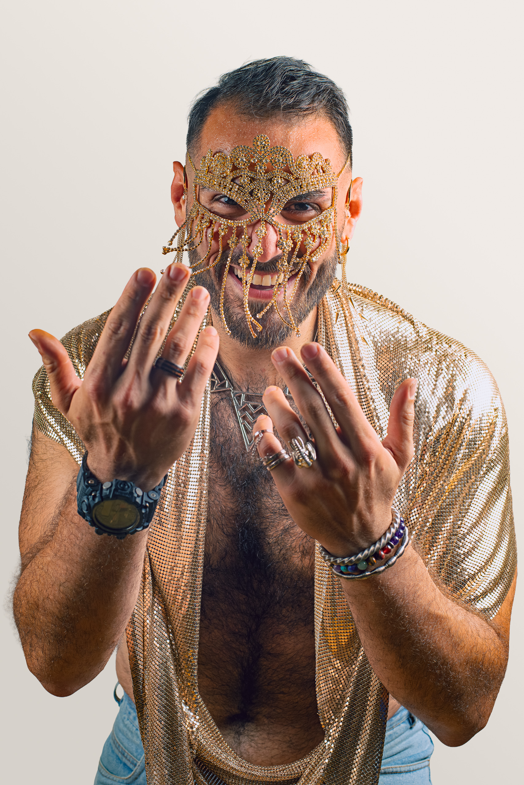 A sitter in a gold beaded mask raises both hands towards the camera.