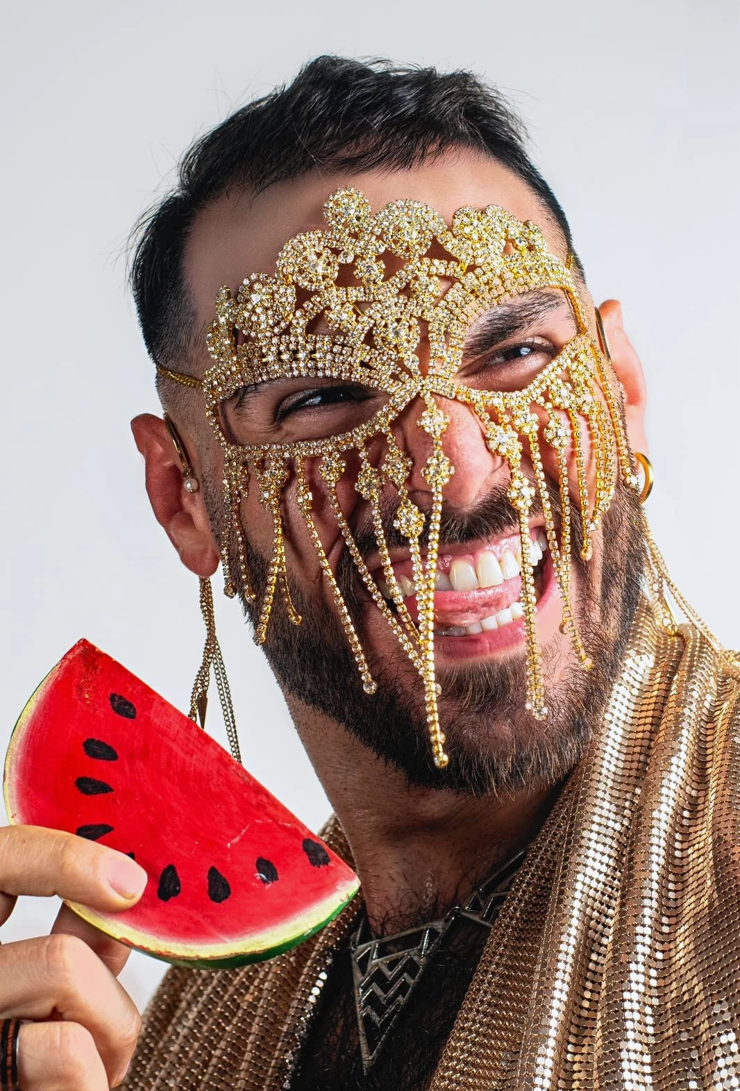 A laughing sitter in a gold beaded mask holds a watermelon-shaped fan close to the face.