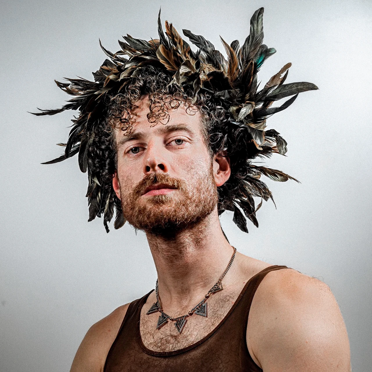 A sitter with curly hair wears a feather crown and a brown mesh top against a light background.