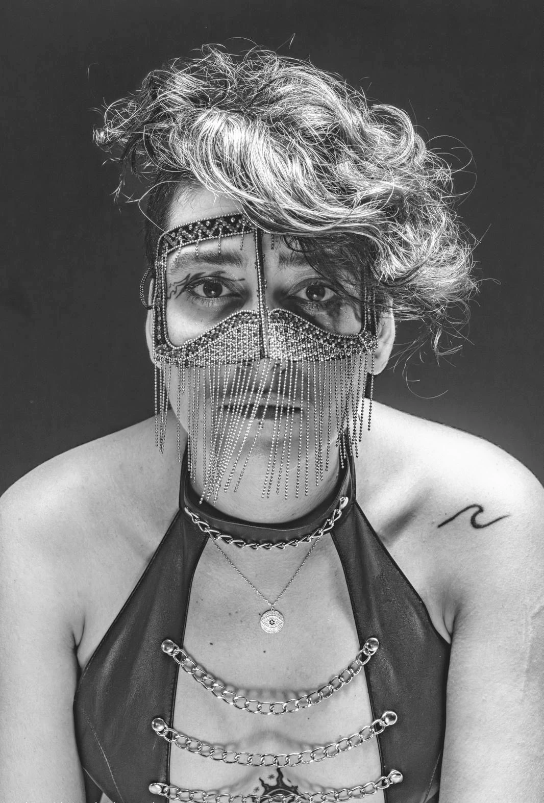 A black-and-white close portrait of a sitter with curled hair, a beaded mask and a black harness.