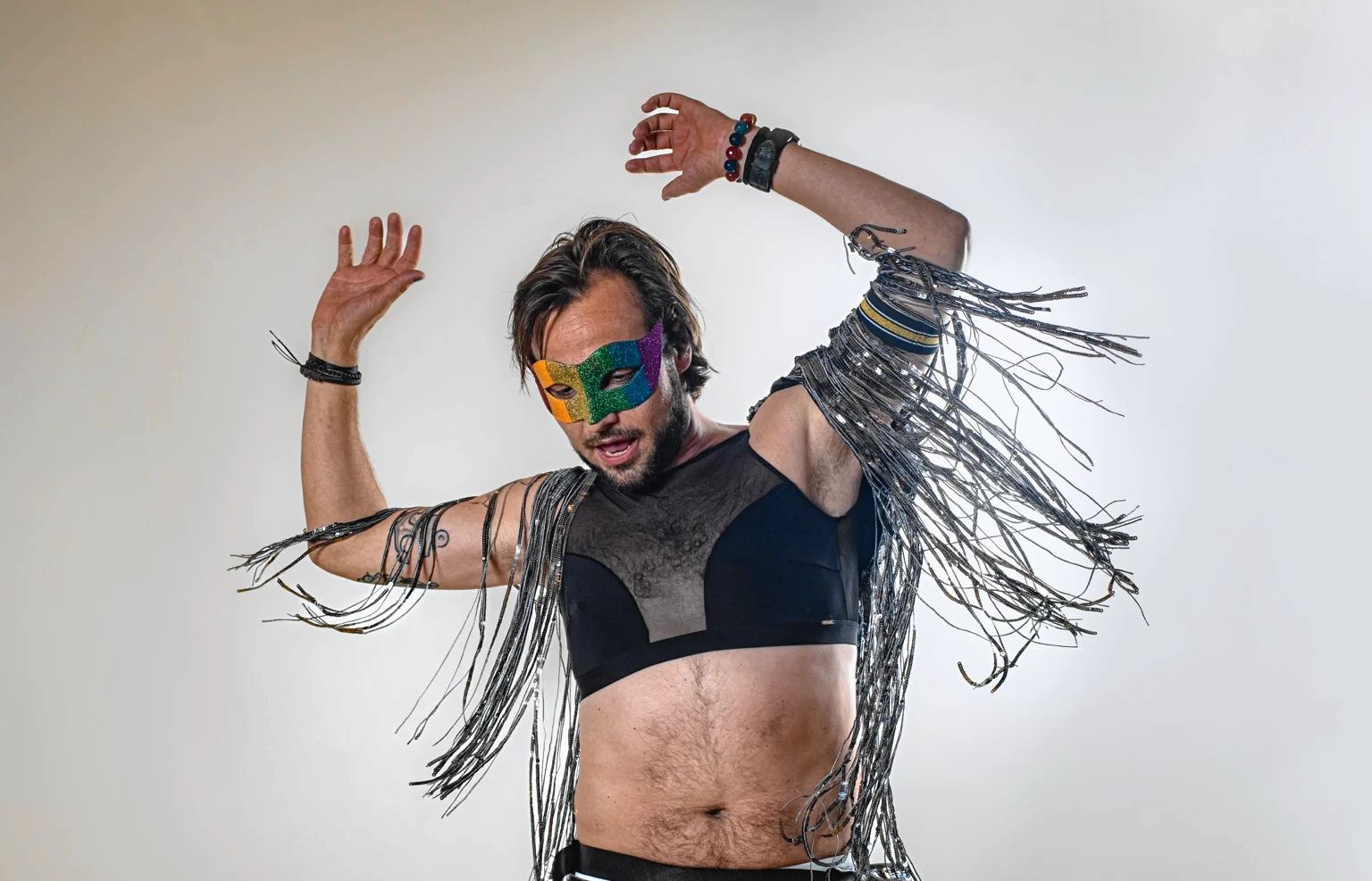 A performer in silver fringe raises both arms, wearing a colourful mask.