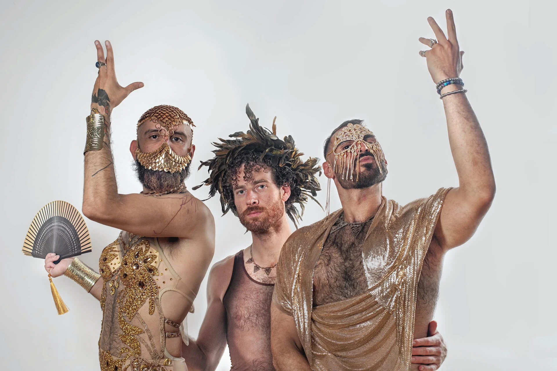 Three sitters pose with raised hands, gold costume, a fan and the feather headpiece against a light wall.