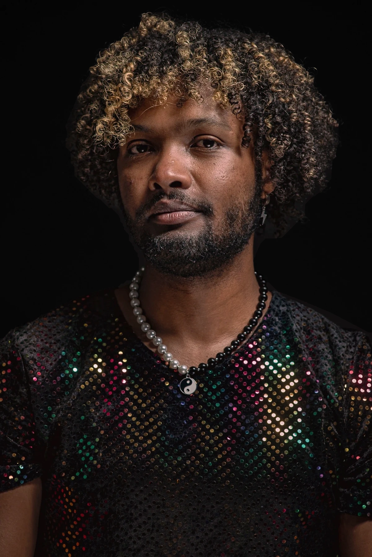 A sitter with highlighted curls wears a multicoloured sequinned top and a beaded necklace against a dark backdrop.