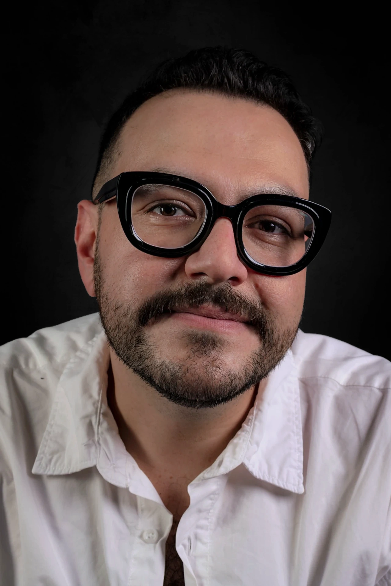 A sitter in thick black glasses and an open-necked white shirt leans towards the camera with a slight smile.