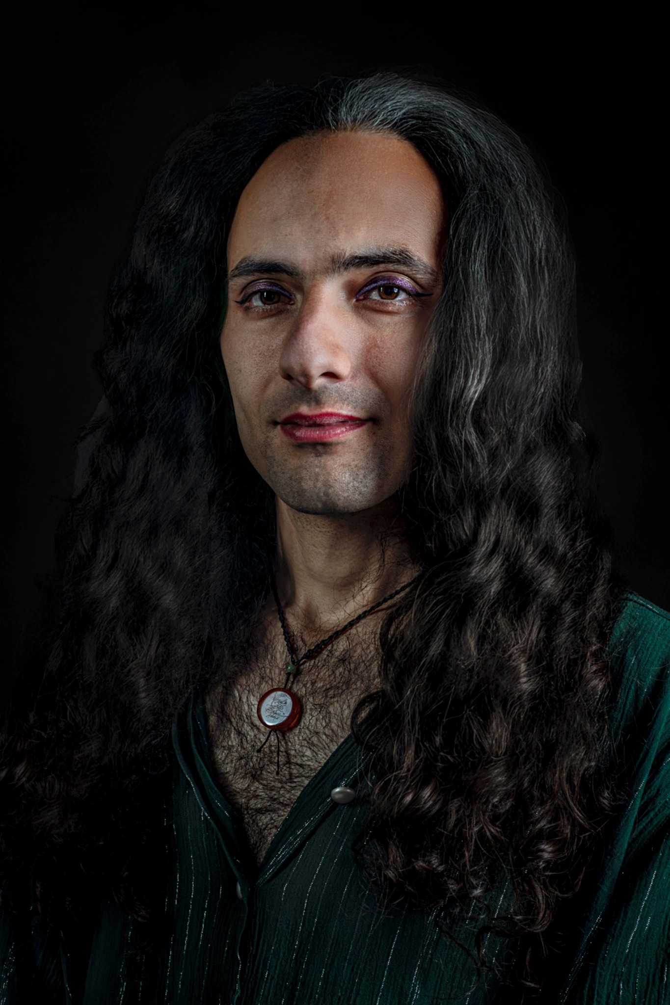 A sitter with long dark hair wears eye makeup, a green shirt and a round pendant.