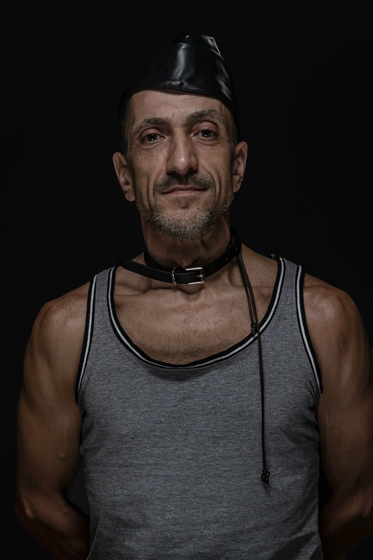 A sitter in a grey tank top, leather cap and collar looks directly at the camera.