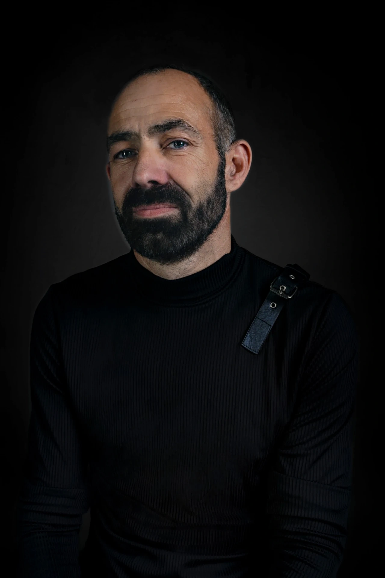 A bearded sitter in a black long-sleeved shirt and shoulder strap looks towards the camera.