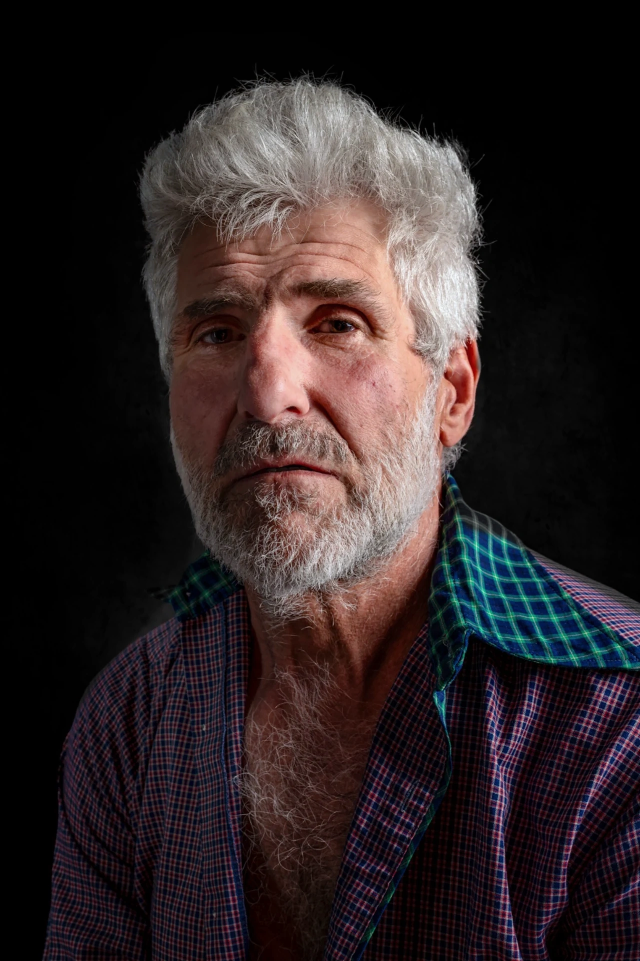 A silver-haired, bearded sitter wears an open-necked purple and green checked shirt against a dark background.