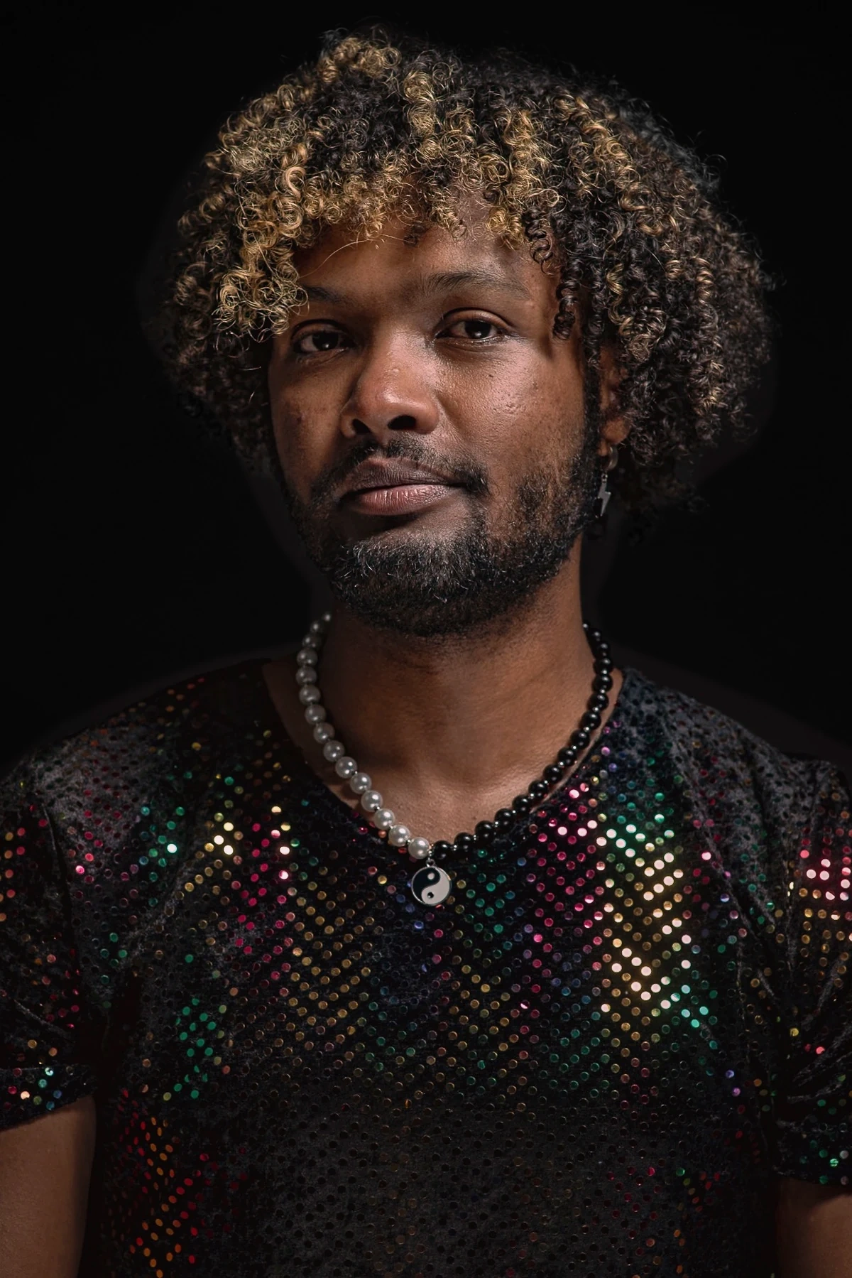 A sitter with highlighted curls wears a multicoloured sequinned top and a beaded necklace against a dark backdrop.