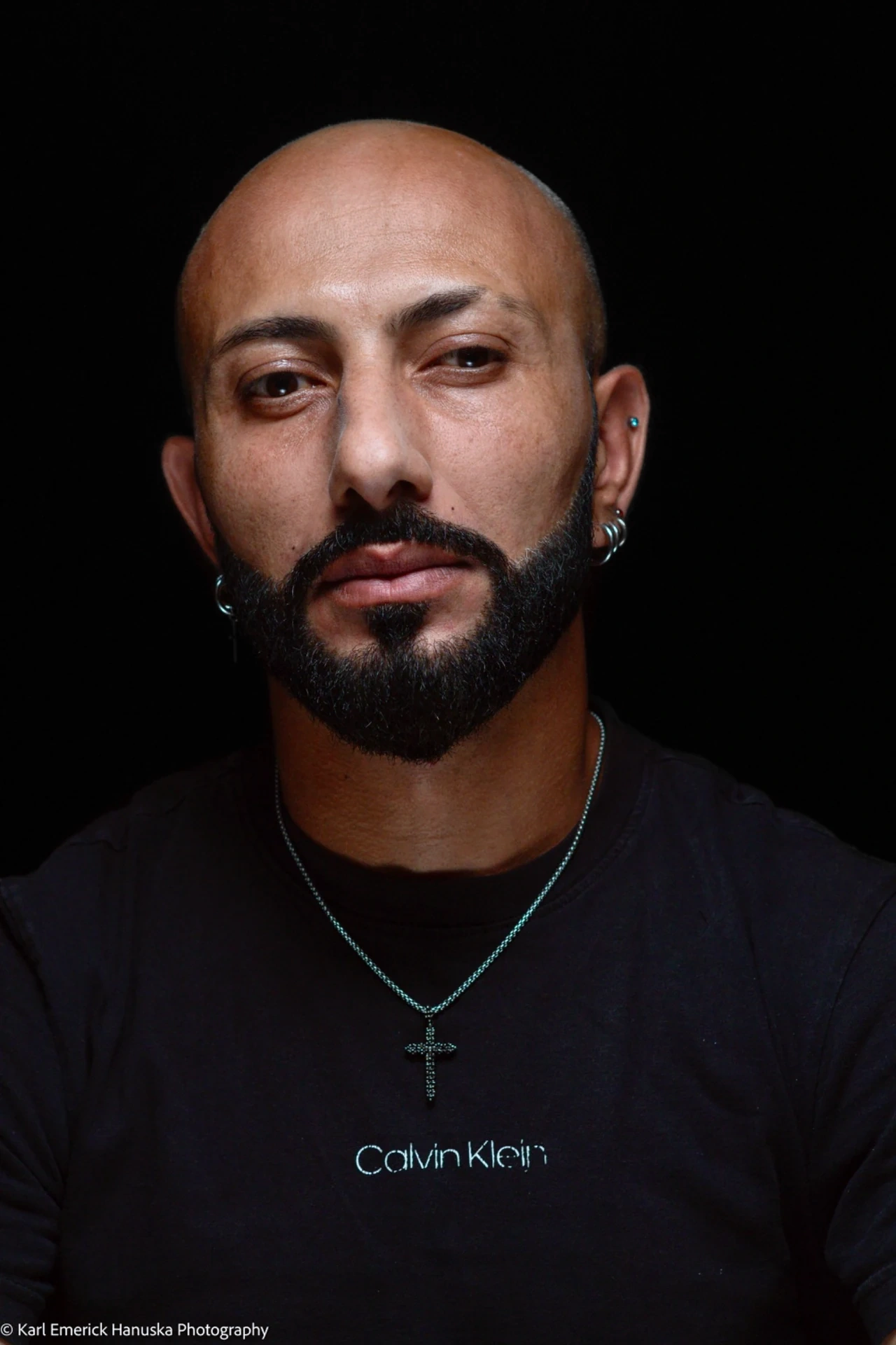 A close portrait of a bearded sitter with a shaved head, earrings and a cross pendant, wearing a black T-shirt.