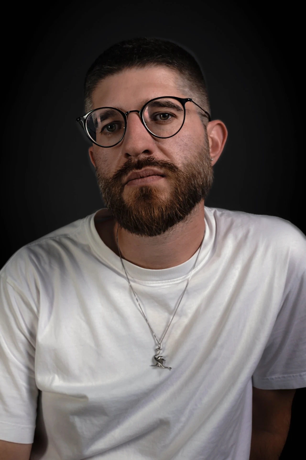 A bearded sitter in round glasses, a white T-shirt and a silver pendant turns slightly towards the camera.