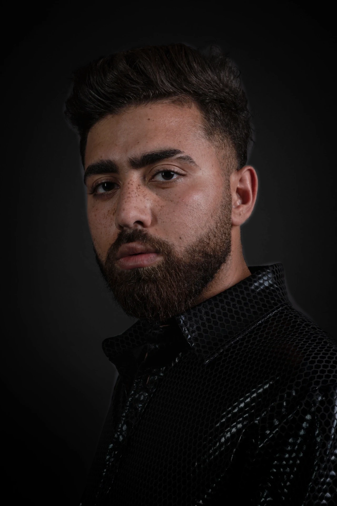 A close portrait of a bearded sitter in a glossy black patterned shirt, turned towards the camera against a charcoal backdrop.