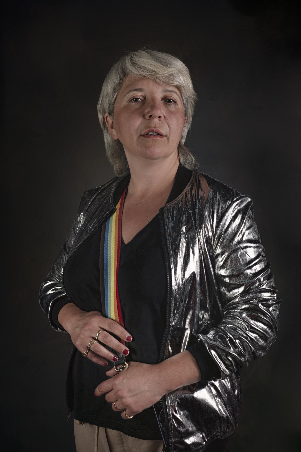 Nata wears a reflective silver jacket over a black top, with a rainbow strap across the chest and hands held together.