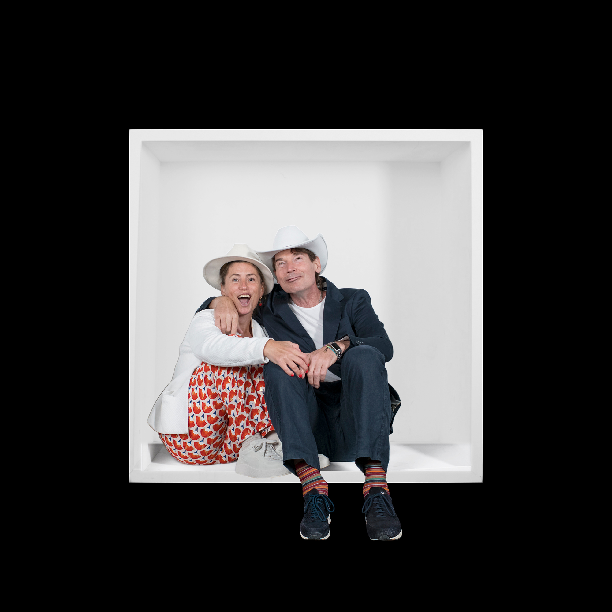 A laughing woman in a white hat sits close beside a man in a white cowboy hat, his arm around her shoulders.