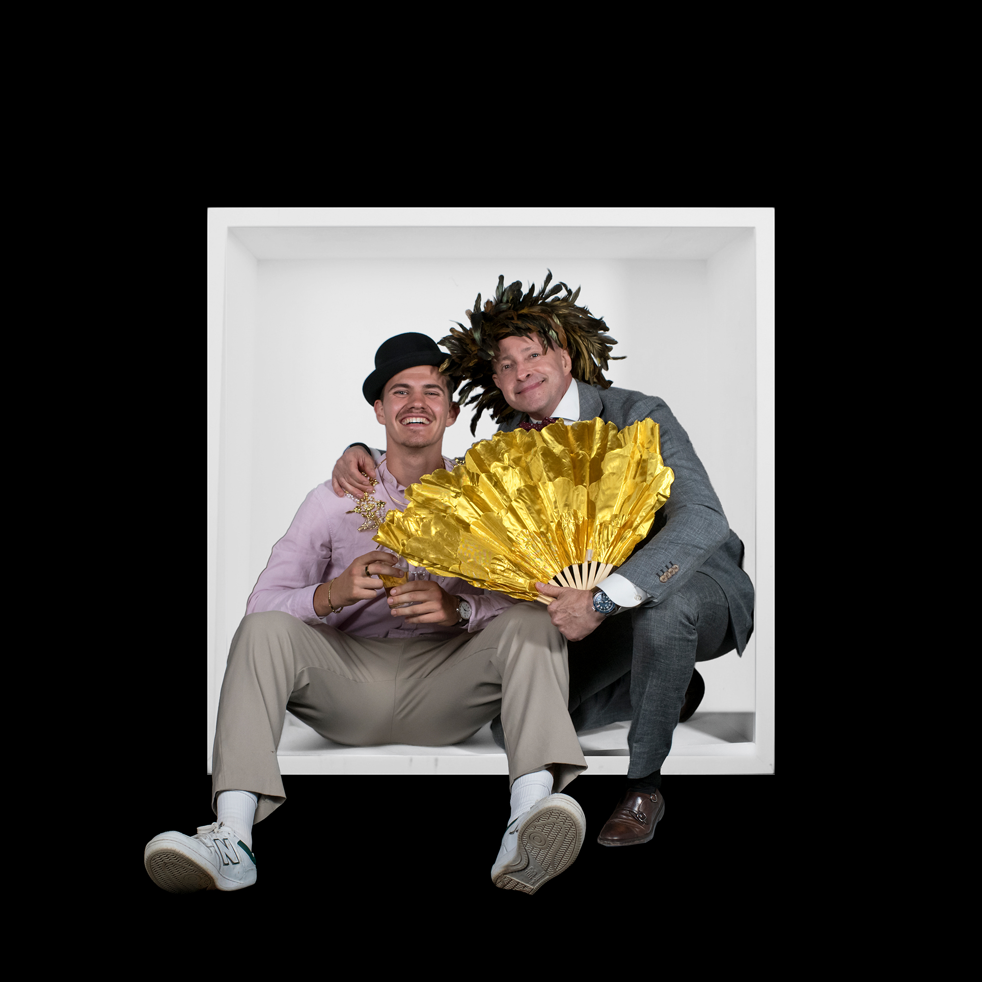 Two smiling men sit close inside the white box, one wearing a tilted black hat and the other a feather headdress while holding a gold fan.