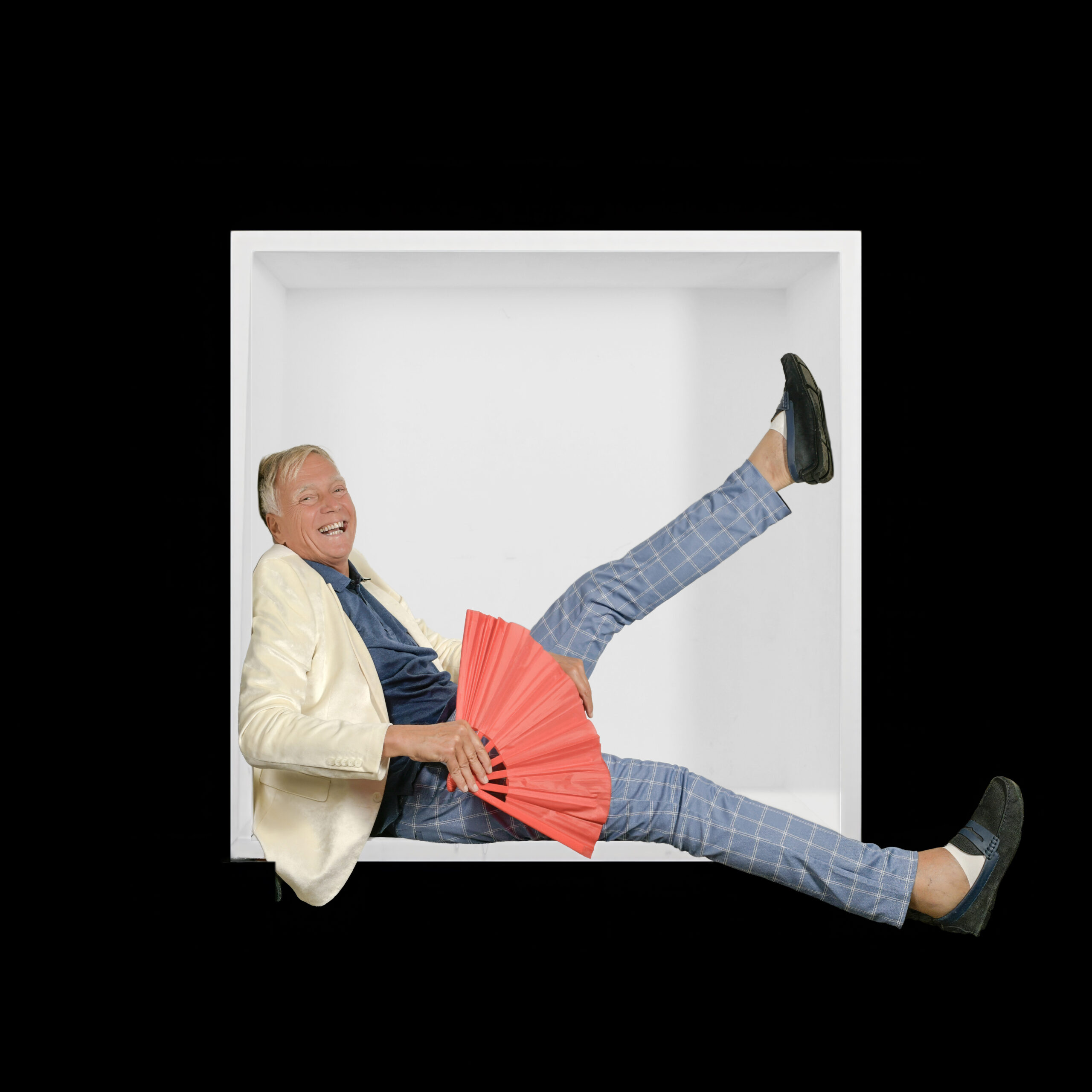A laughing older man in a pale jacket holds a red fan and kicks one leg high across the white box.