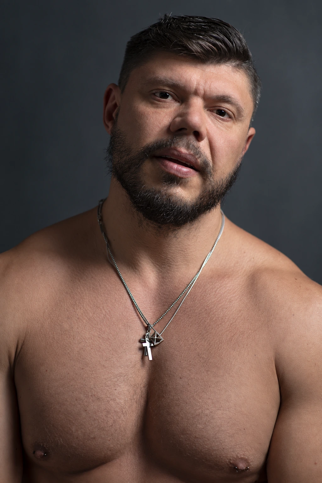 A bare-chested bearded sitter with two necklaces looks directly at the camera against a softly lit dark grey background.