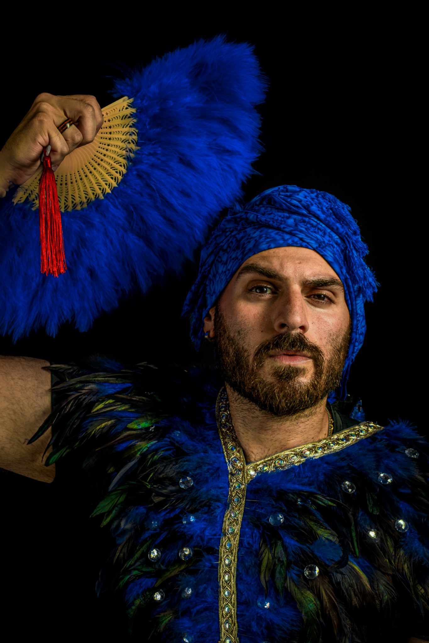 A sitter in a blue turban and feathered costume holds a blue fan above the head.