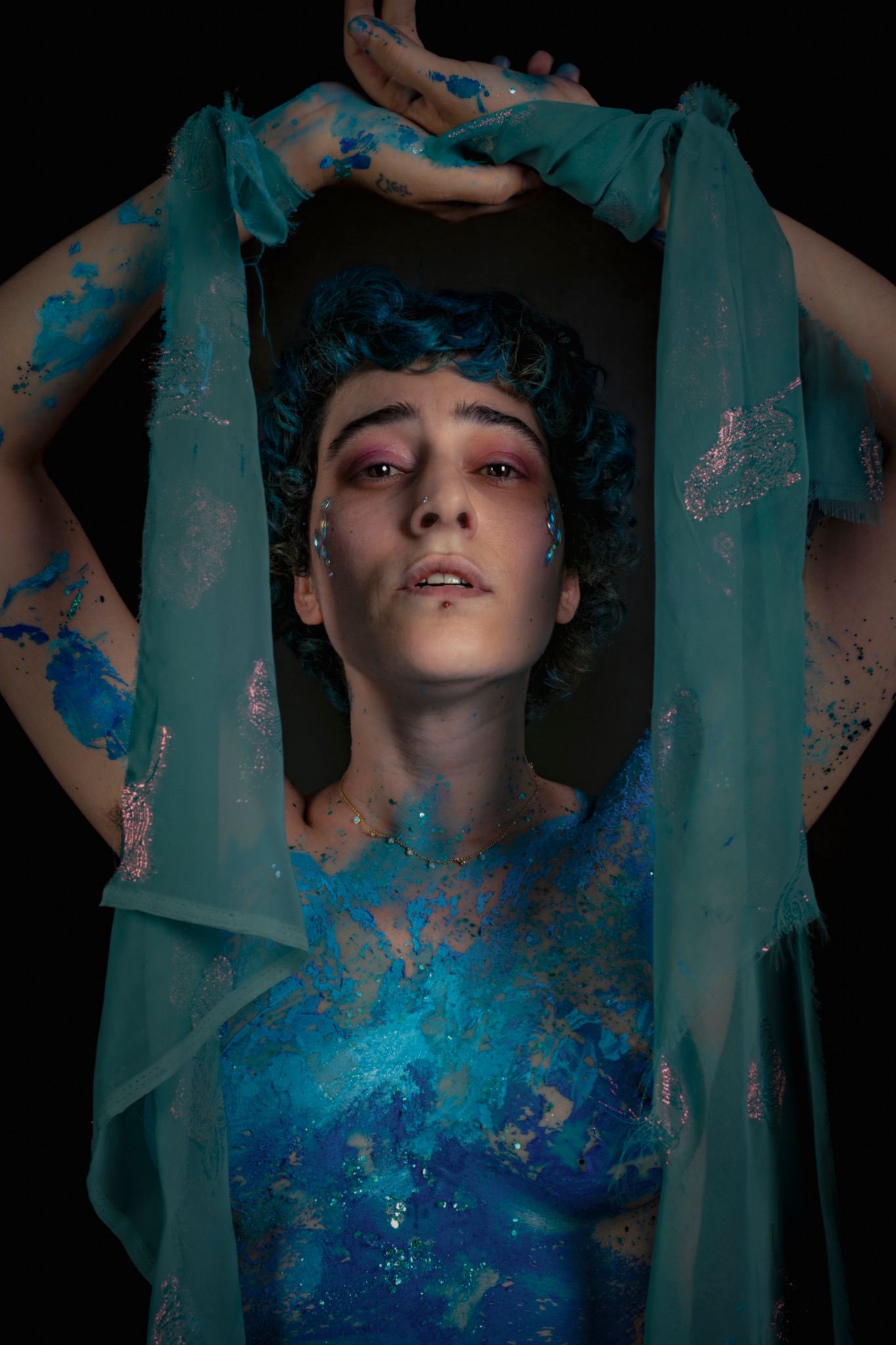 A sitter with blue body paint raises both arms around sheer turquoise fabric.