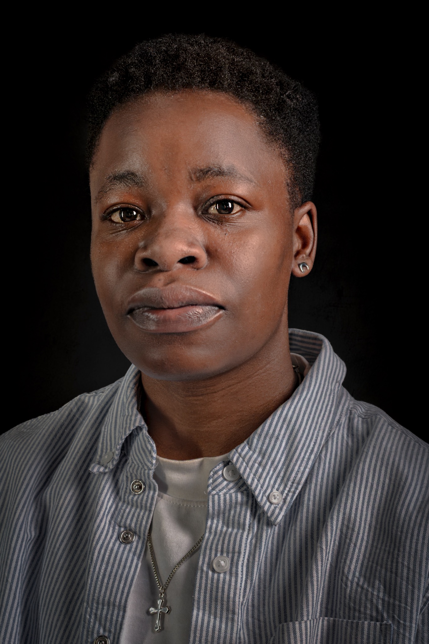 A close portrait of a sitter with short natural hair, a striped shirt and a silver cross necklace.