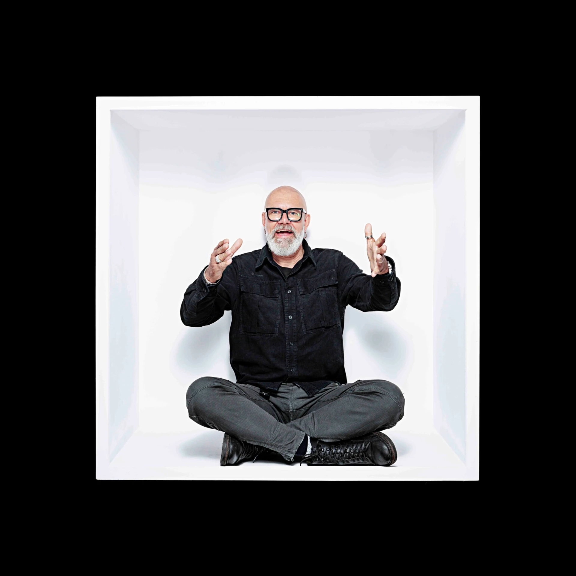 A bearded sitter with glasses sits cross-legged inside a white box, hands raised.