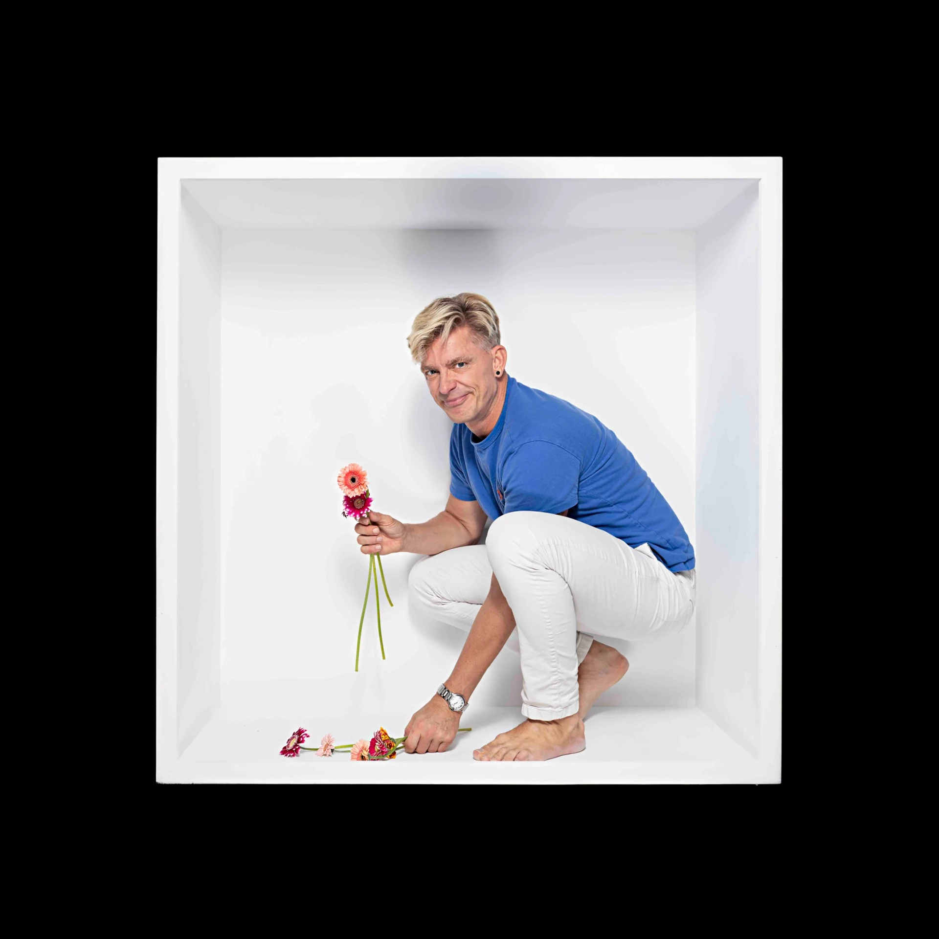 A sitter in a blue shirt crouches inside a white box holding pink flowers.