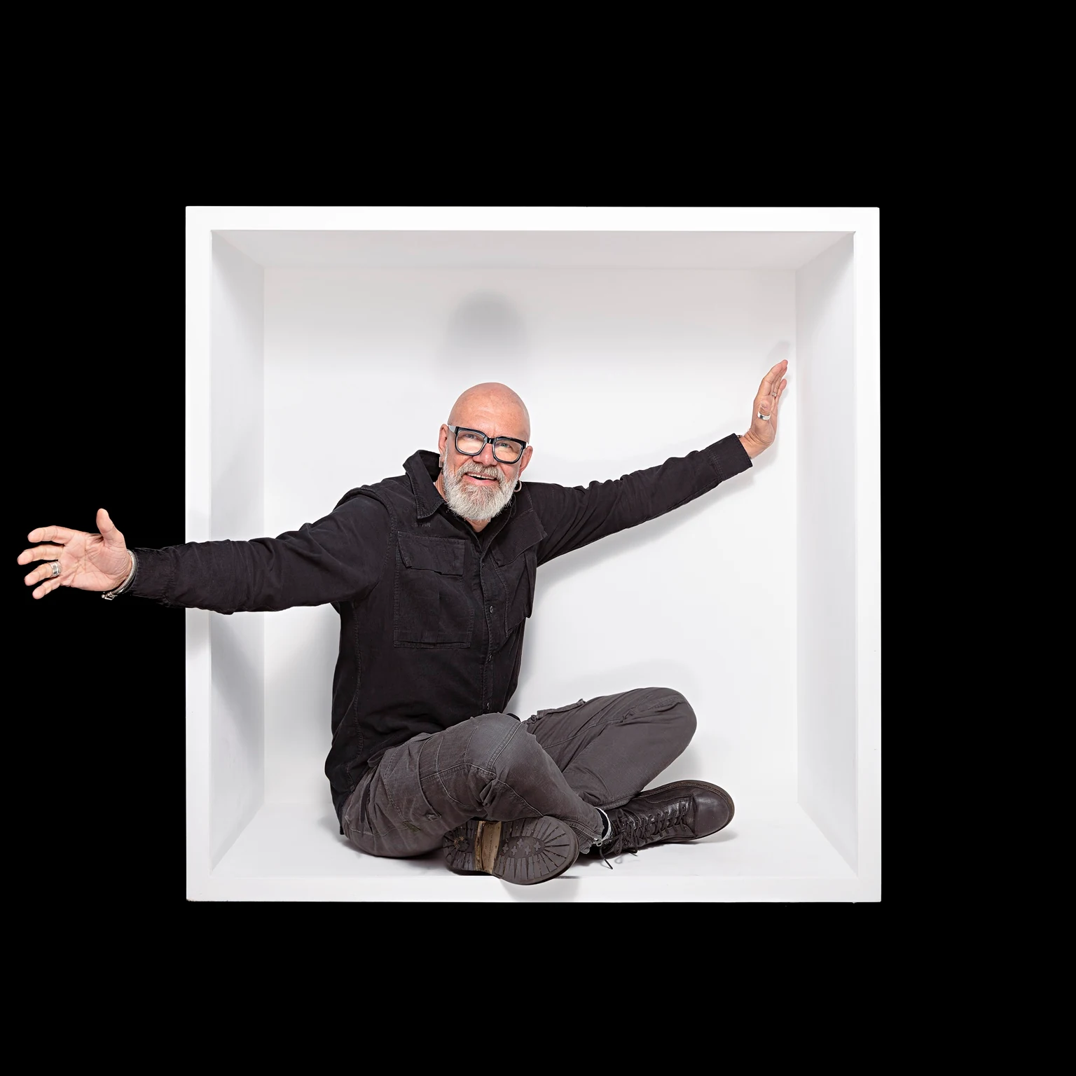 A bearded sitter with glasses sits cross-legged in a white box, extending both arms into an open gesture.
