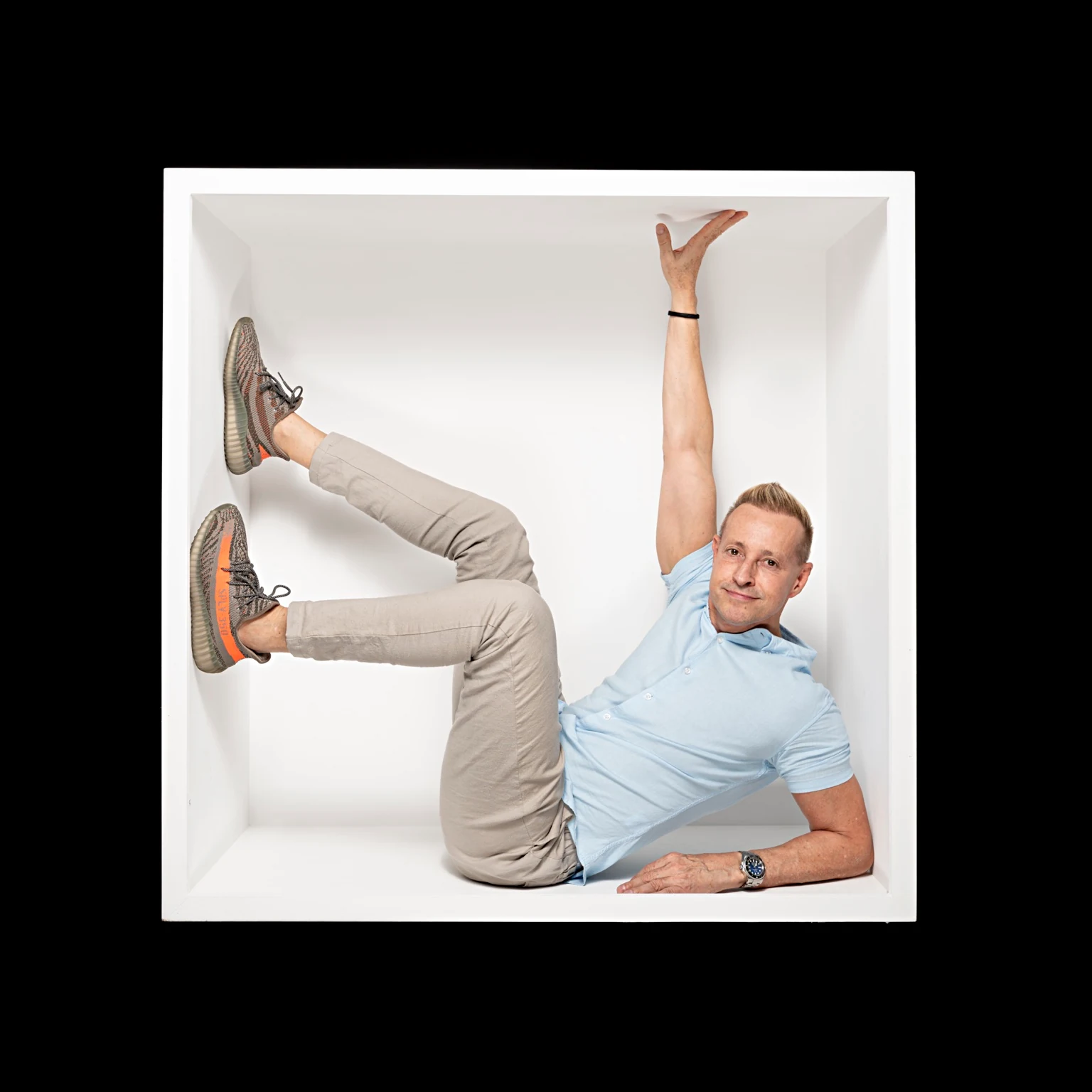 A sitter in a pale blue polo shirt reclines diagonally inside a white box, with raised legs and one arm reaching upward.
