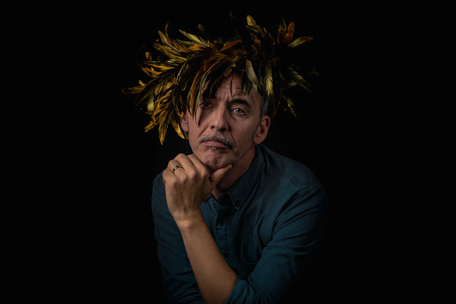 Een portret in een blauw shirt met een kroon van gouden veren en een hand onder de kin.