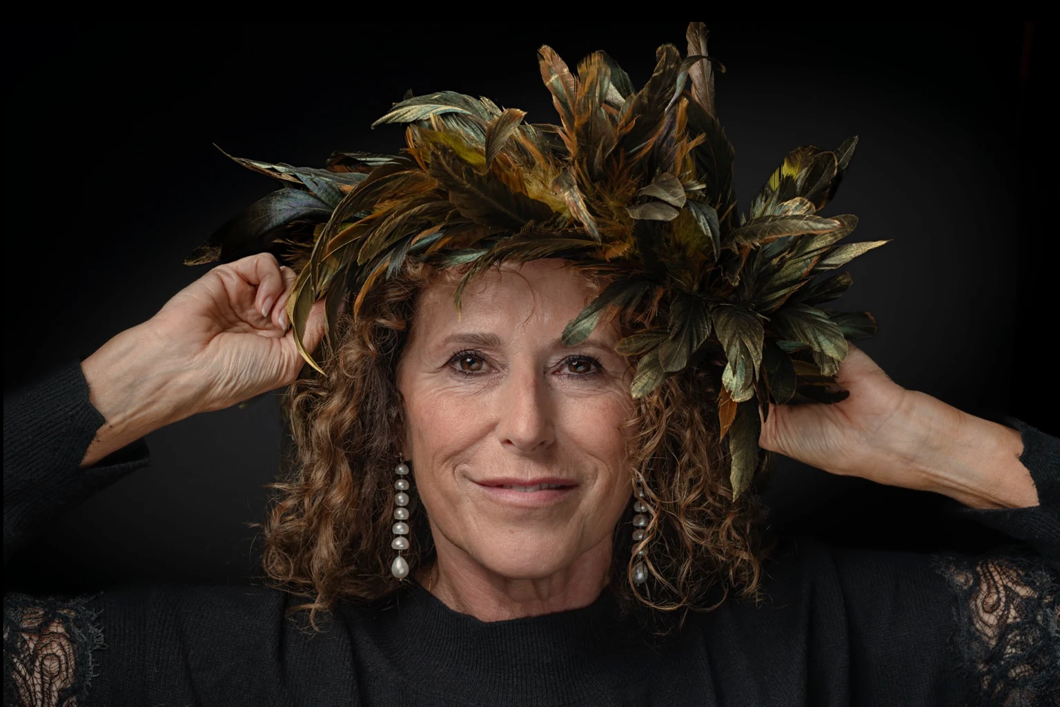 A curly-haired sitter with pearl earrings raises both hands to a golden feather crown.
