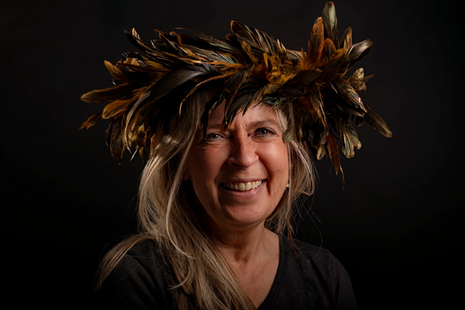 A smiling sitter with long fair hair wears a golden feather crown against a black background.