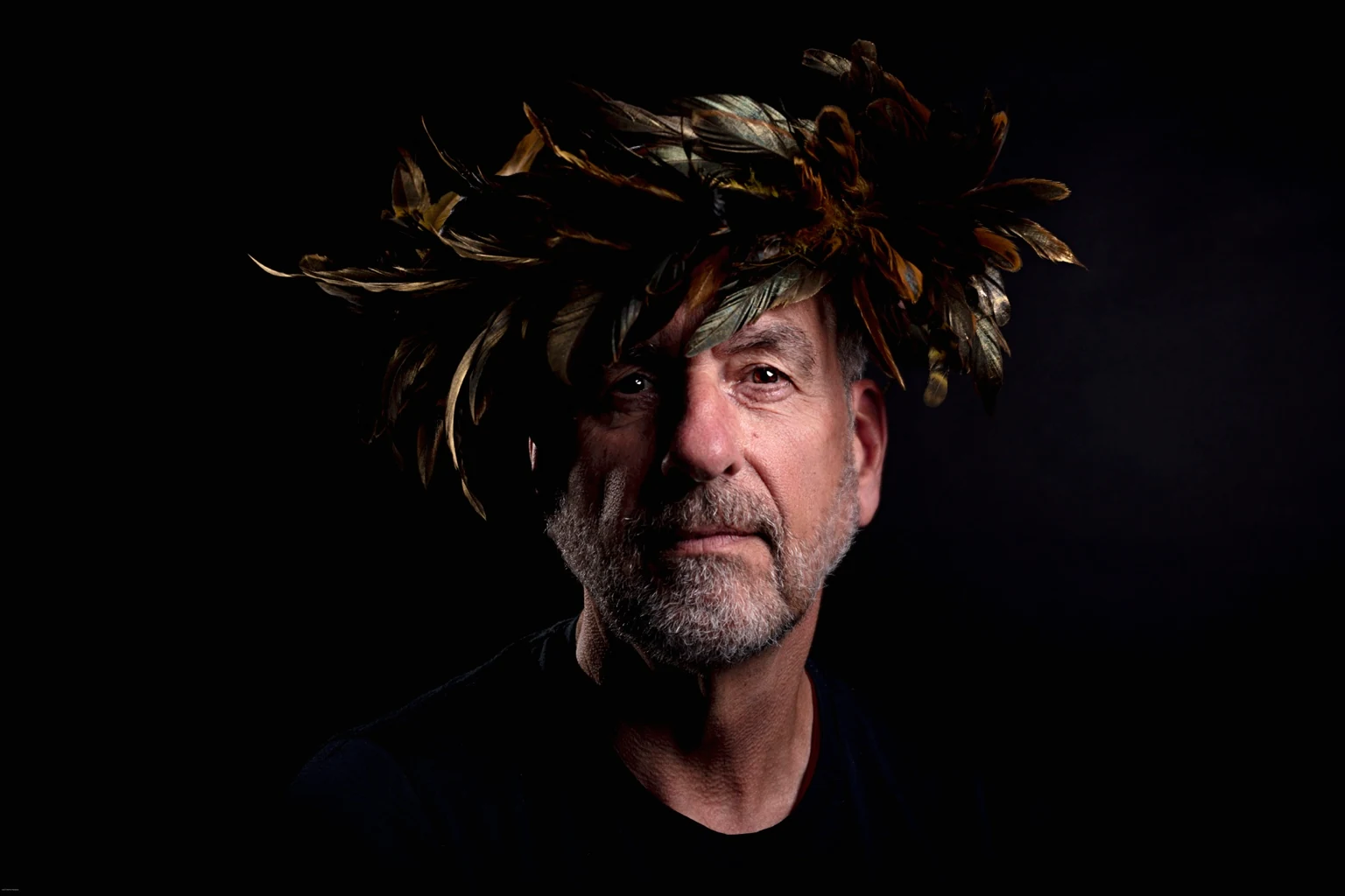A grey-bearded sitter in a black shirt wears a golden feather crown, with one side of the face in shadow.
