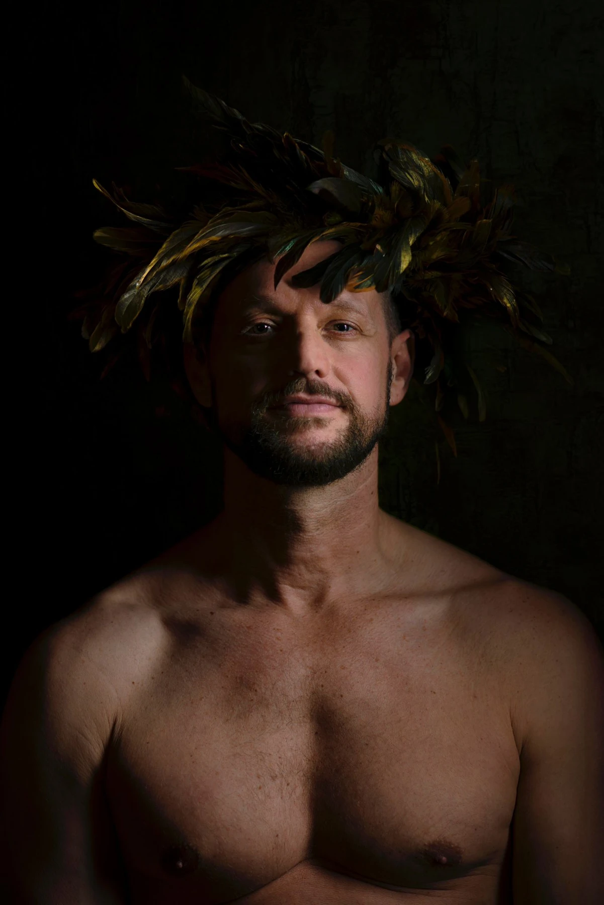A bare-chested sitter looks directly at the camera beneath a crown of golden feathers, with one side of the face in shadow.