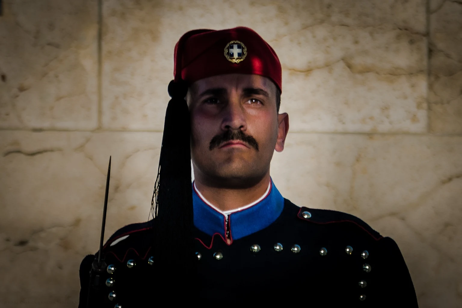 A guard in a dark ceremonial uniform and red cap stands against a warmly lit marble wall.