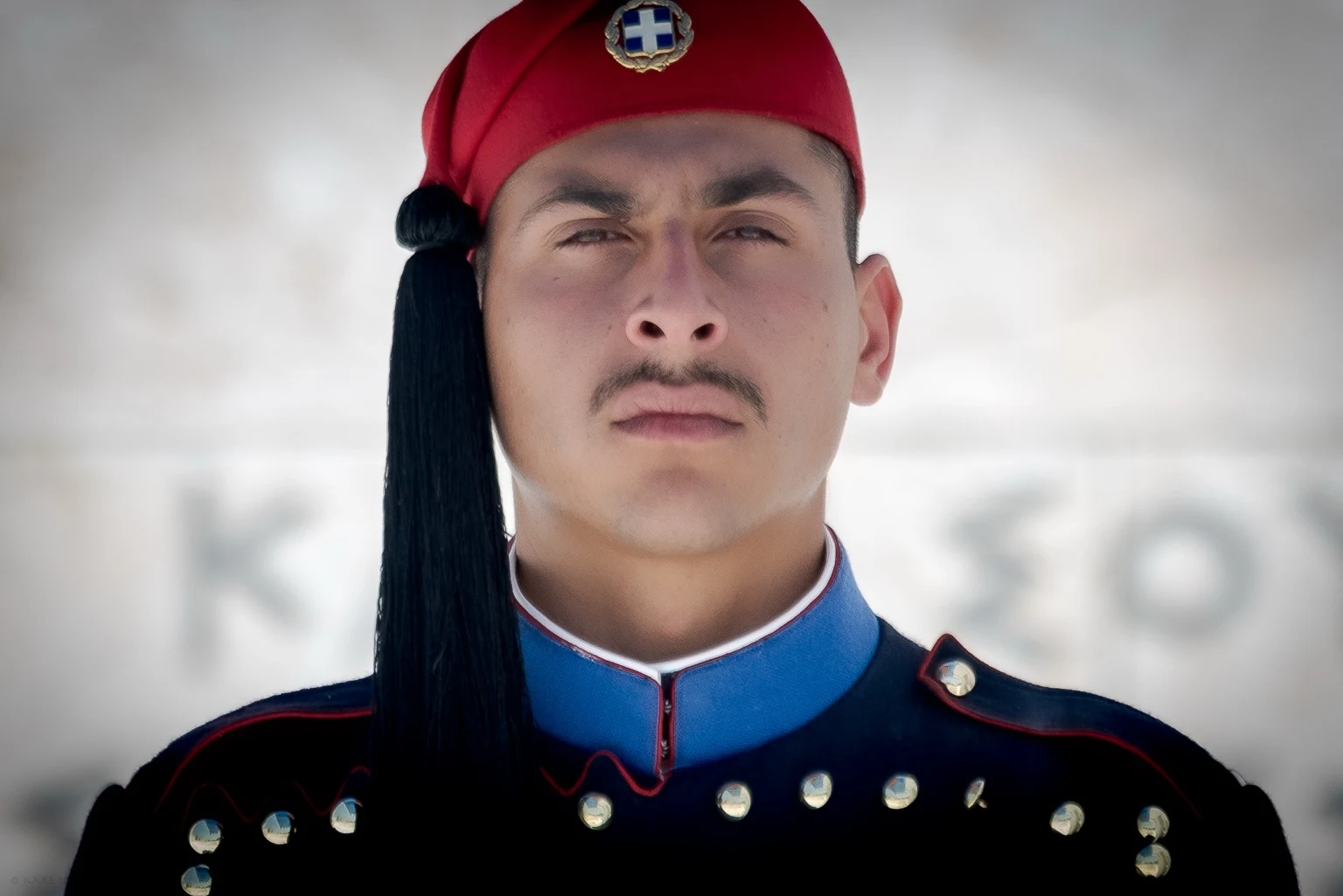 A close portrait of a guard in a dark ceremonial uniform, with a red cap and long black tassel.
