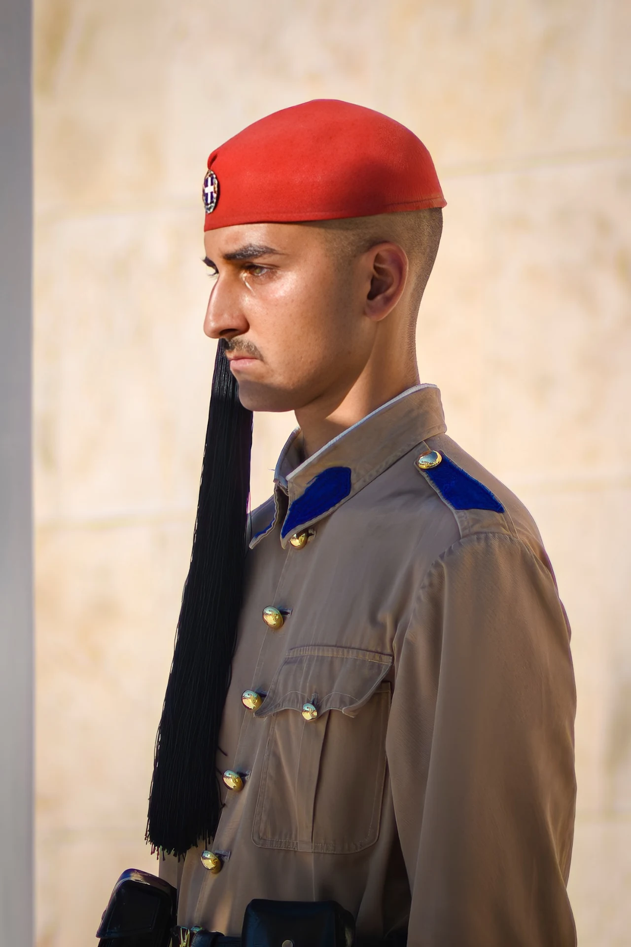 A guard in a red cap and tan uniform stands in profile against a softly lit stone background.